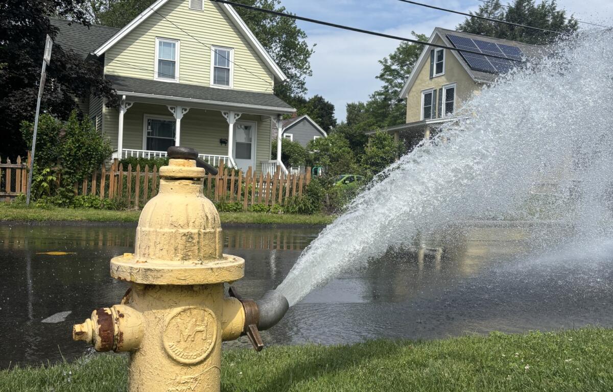 Yellow fire hydrant in a grassy verge spraying a large arc of water across a suburban street with houses in the background under a blue sky.
