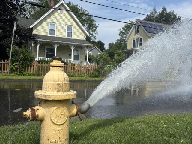 Yellow fire hydrant in a grassy verge spraying a large arc of water across a suburban street with houses in the background under a blue sky.