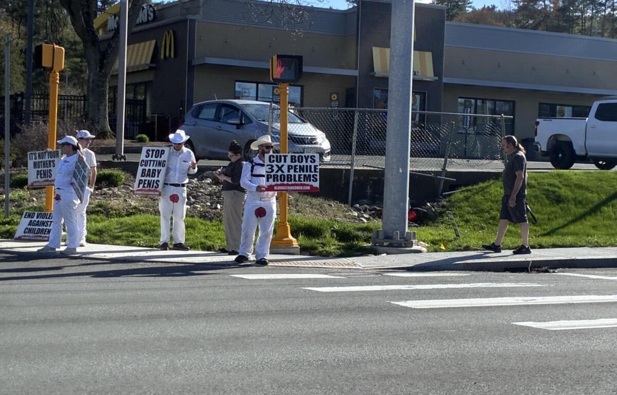 Group of protesters in white clothing holding signs at a street corner with a fast-food restaurant in the background (McDonald's).