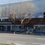Construction workers install large blue letters on the storefront facade; a lift on the left and a yellow construction vehicle on the right.