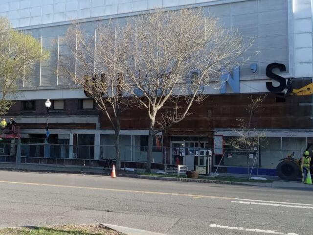 Construction workers install large blue letters on the storefront facade; a lift on the left and a yellow construction vehicle on the right.