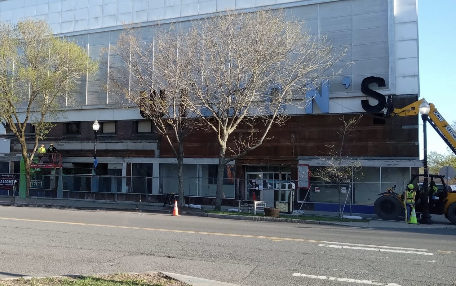 Construction workers install large blue letters on the storefront facade; a lift on the left and a yellow construction vehicle on the right.