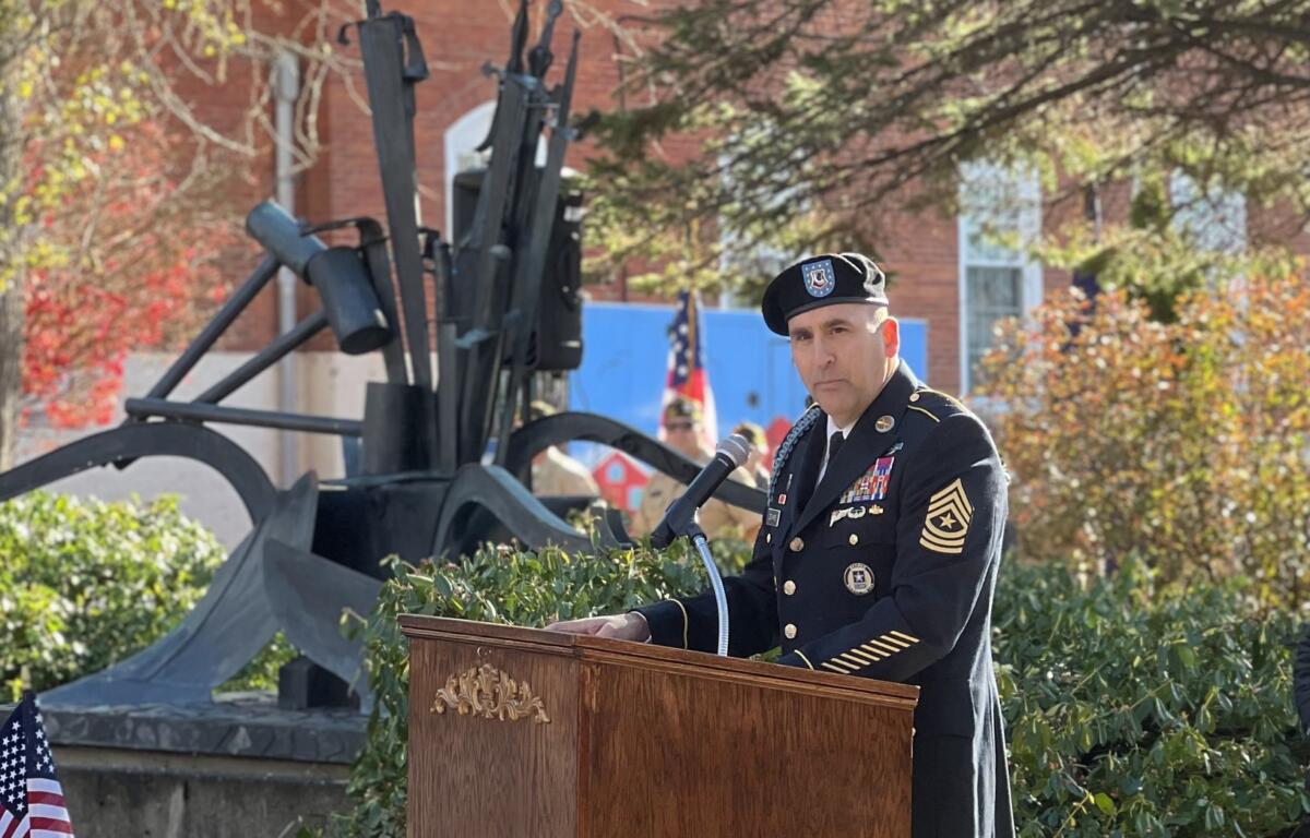 Military officer in dress uniform delivering a speech at an outdoor ceremony, with a cannon display and flags in the background.