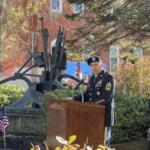 Military officer in dress uniform delivering a speech at an outdoor ceremony, with a cannon display and flags in the background.