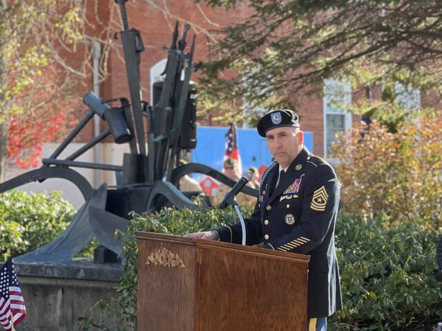 Military officer in dress uniform delivering a speech at an outdoor ceremony, with a cannon display and flags in the background.