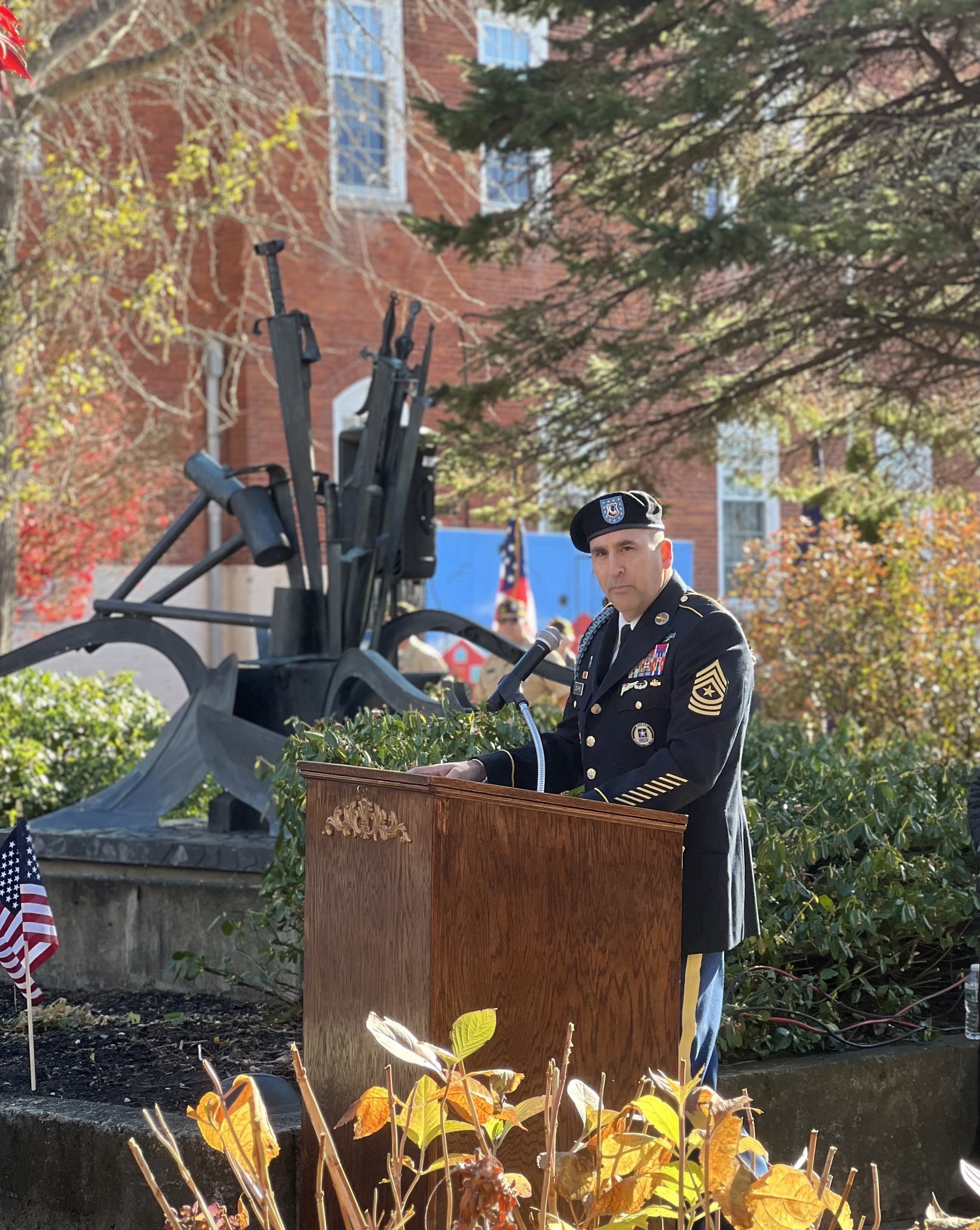 Military officer in dress uniform delivering a speech at an outdoor ceremony, with a cannon display and flags in the background.