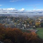 Aerial view of a fall-colored town with orange and yellow trees and fog drifting over rooftops under a blue sky.