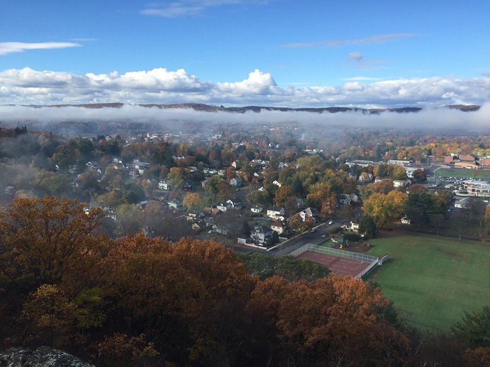 Aerial view of a fall-colored town with orange and yellow trees and fog drifting over rooftops under a blue sky.