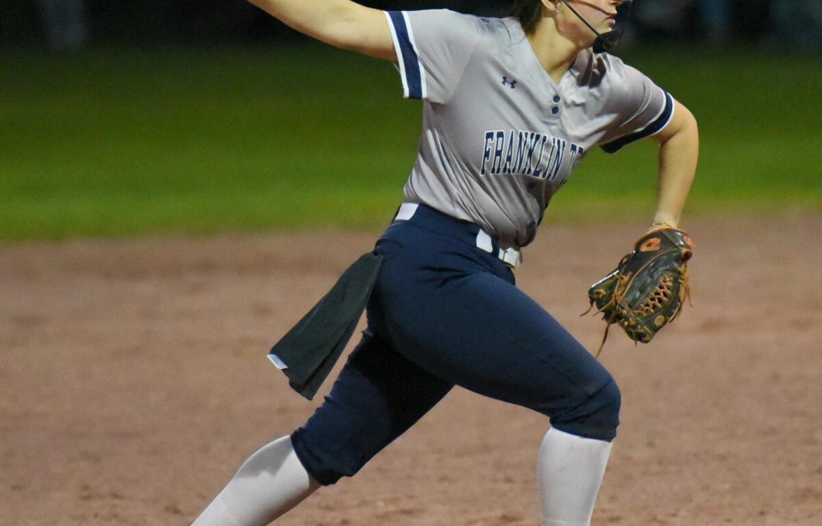 Female softball pitcher in a gray Franklin uniform releasing a neon yellow ball on a dirt mound at night, wearing a protective mask and cleats.