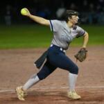 Female softball pitcher in a gray Franklin uniform releasing a neon yellow ball on a dirt mound at night, wearing a protective mask and cleats.