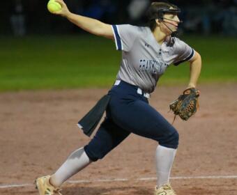 Female softball pitcher in a gray Franklin uniform releasing a neon yellow ball on a dirt mound at night, wearing a protective mask and cleats.