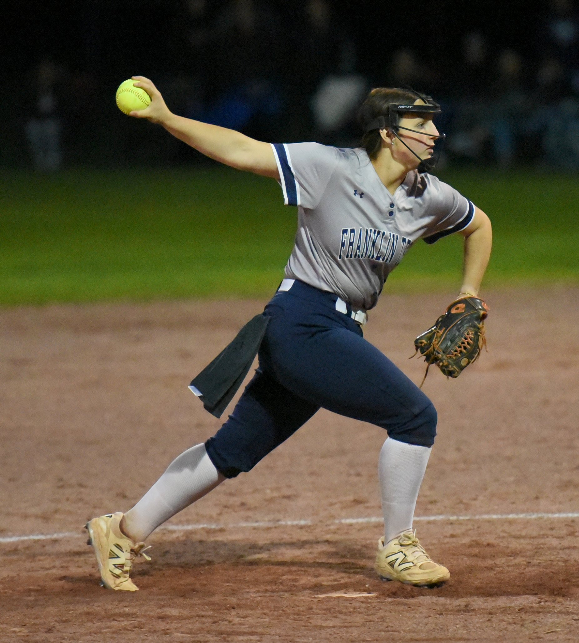 Female softball pitcher in a gray Franklin uniform releasing a neon yellow ball on a dirt mound at night, wearing a protective mask and cleats.