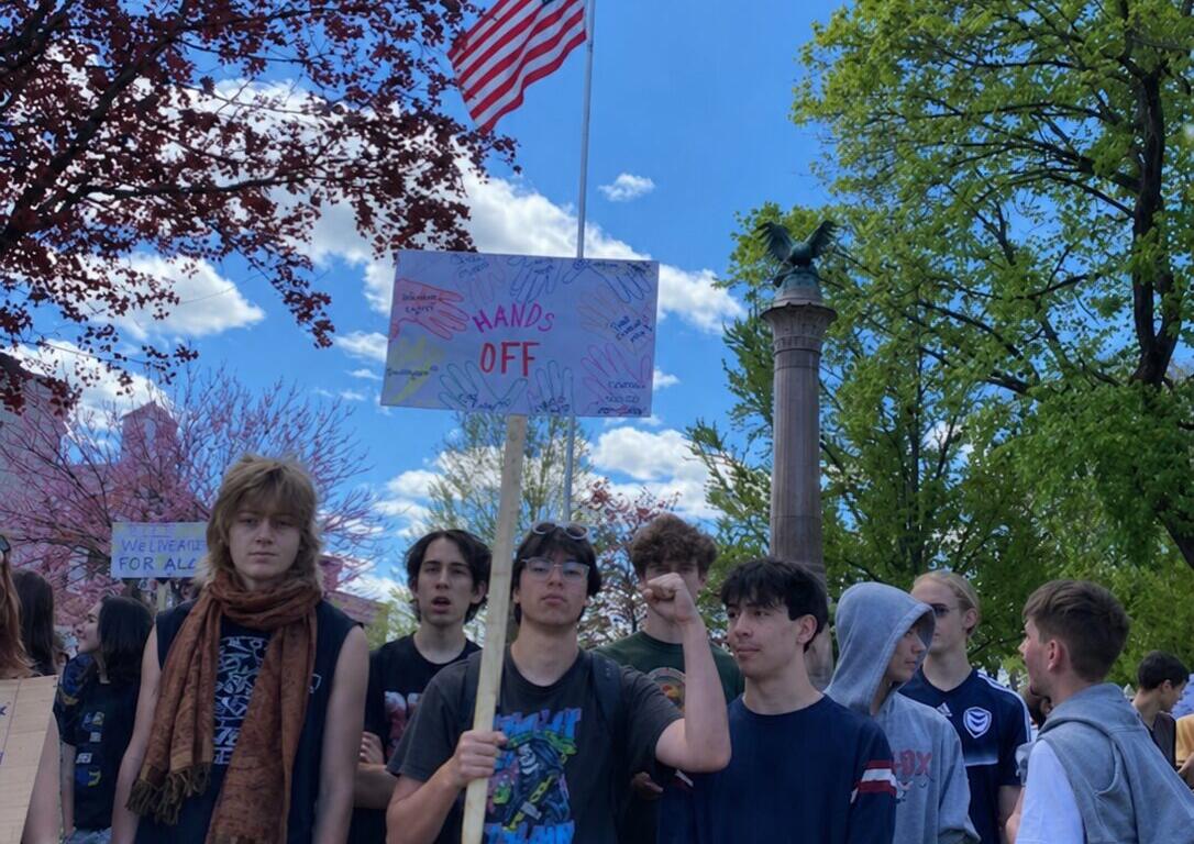 A group of young people gathered outdoors at a protest, one person holds a sign reading 'HANDS OFF' under a waving American flag against a bright blue sky.