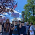 A group of young people gathered outdoors at a protest, one person holds a sign reading 'HANDS OFF' under a waving American flag against a bright blue sky.