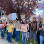 Group of diverse young protesters holding handmade signs on a city sidewalk with red-leaf trees overhead, signaling a rights demonstration.