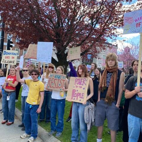 Group of diverse young protesters holding handmade signs on a city sidewalk with red-leaf trees overhead, signaling a rights demonstration.