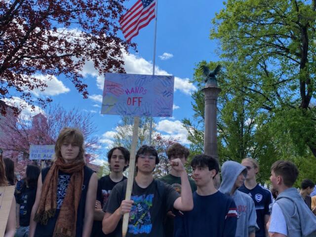 A group of young people gathered outdoors at a protest, one person holds a sign reading 'HANDS OFF' under a waving American flag against a bright blue sky.