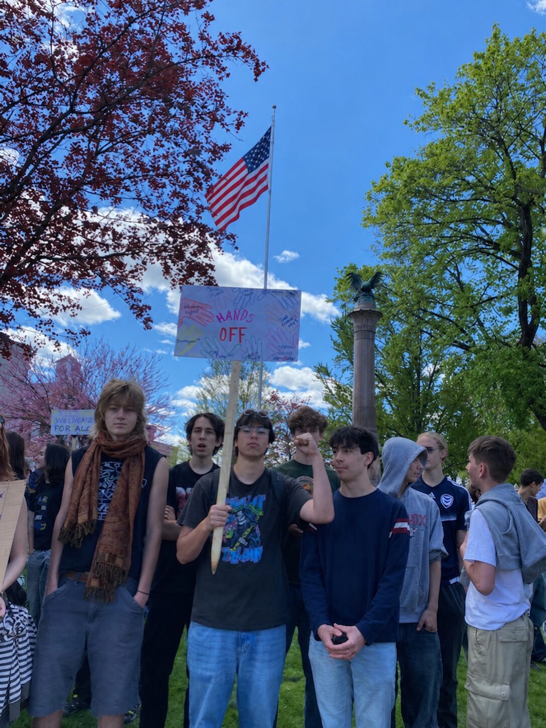 A group of young people gathered outdoors at a protest, one person holds a sign reading 'HANDS OFF' under a waving American flag against a bright blue sky.