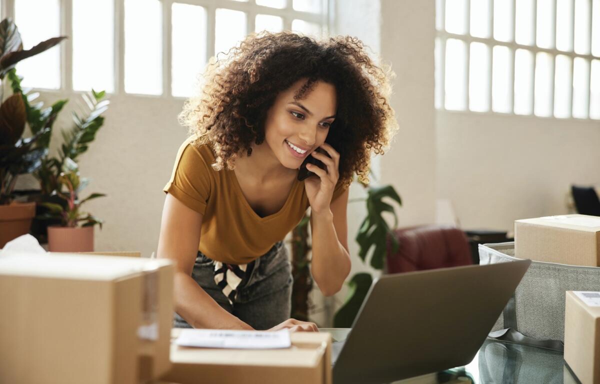 Smiling woman with curly hair on a phone call, leaning over a laptop amid packing boxes in a bright office environment.