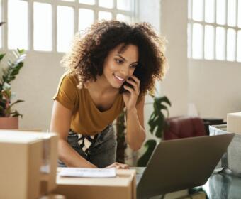 Smiling woman with curly hair on a phone call, leaning over a laptop amid packing boxes in a bright office environment.