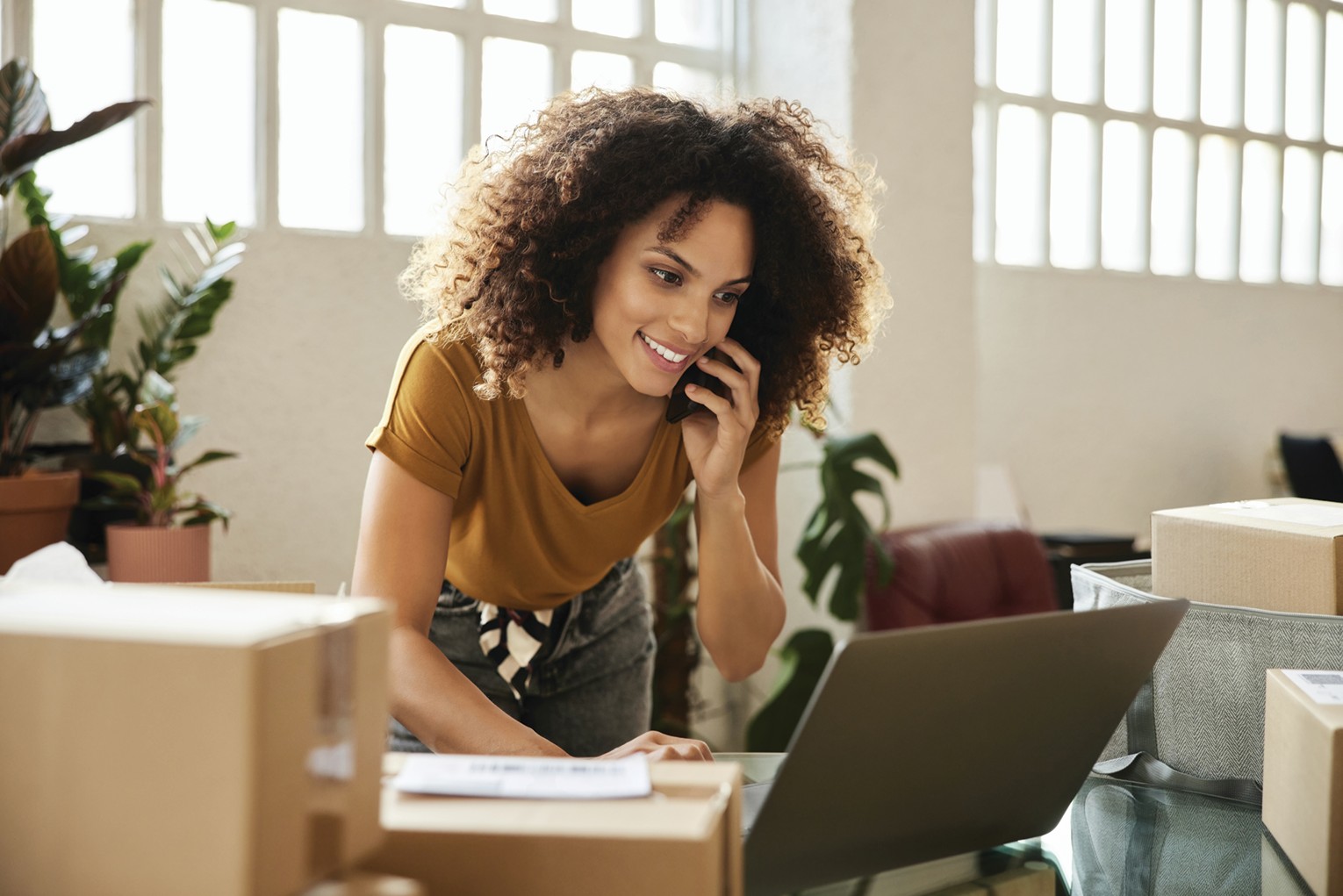 Smiling woman with curly hair on a phone call, leaning over a laptop amid packing boxes in a bright office environment.