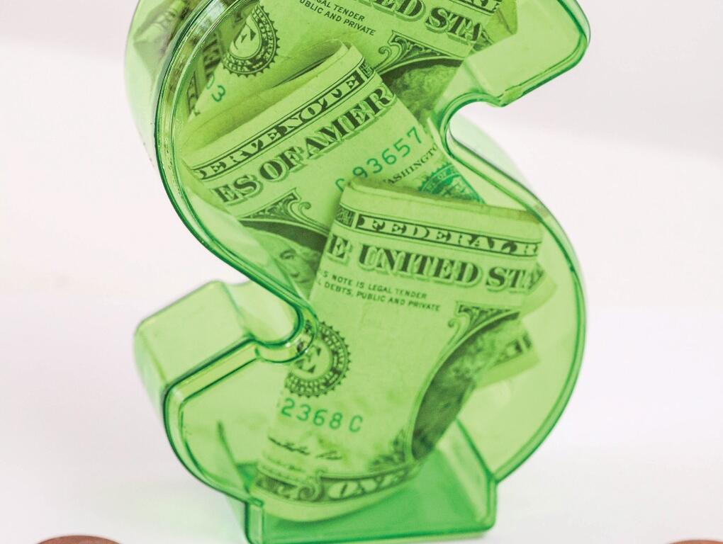 Green transparent dollar-sign piggy bank filled with cash, sitting on a white surface with the text 'What’s your credit score?' nearby and a few coins around it, emphasizing money and credit.