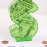 Green transparent dollar-sign piggy bank filled with cash, sitting on a white surface with the text 'What’s your credit score?' nearby and a few coins around it, emphasizing money and credit.