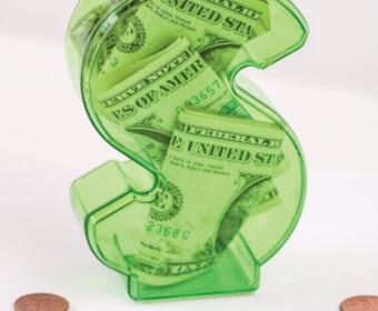 Green transparent dollar-sign piggy bank filled with cash, sitting on a white surface with the text 'What’s your credit score?' nearby and a few coins around it, emphasizing money and credit.
