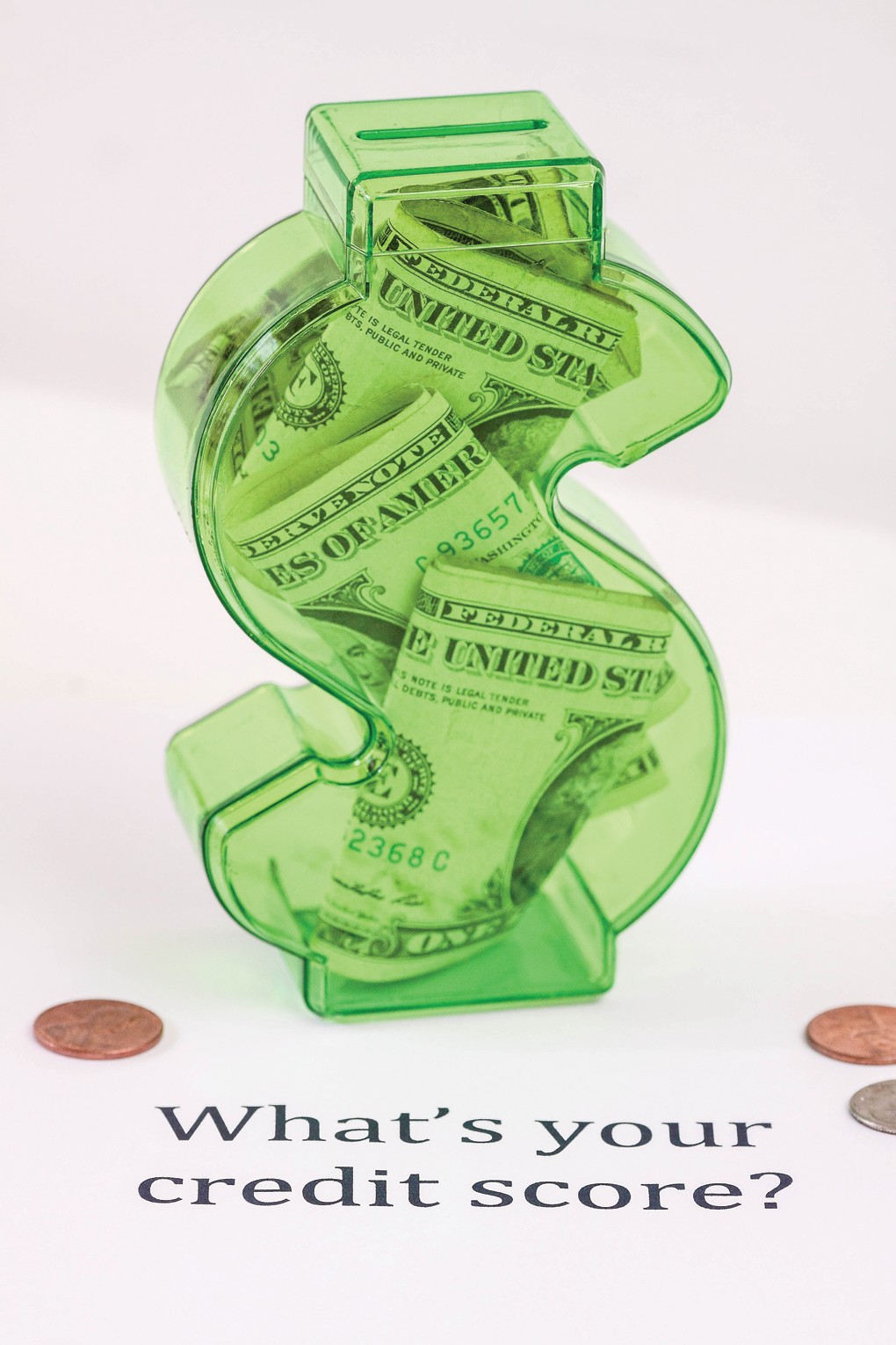 Green transparent dollar-sign piggy bank filled with cash, sitting on a white surface with the text 'What’s your credit score?' nearby and a few coins around it, emphasizing money and credit.