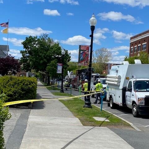 Sidewalk scene with yellow caution tape, utility trucks and workers in safety vests on a sunny day.
