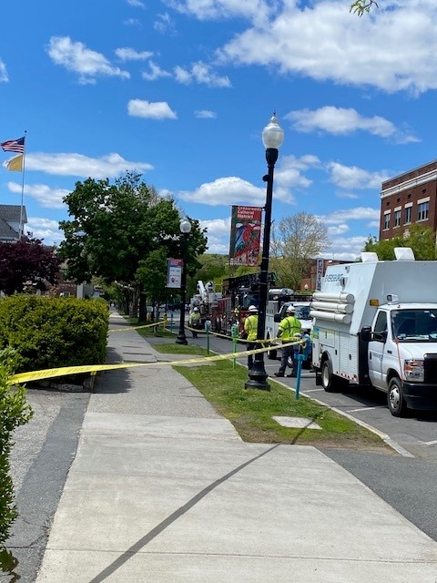 Sidewalk scene with yellow caution tape, utility trucks and workers in safety vests on a sunny day.