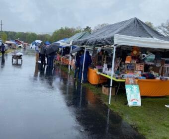 Rainy outdoor market with stalls under tents along a wet, reflective walkway; shoppers carry umbrellas as they browse vendors.