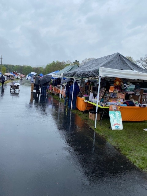 Rainy outdoor market with stalls under tents along a wet, reflective walkway; shoppers carry umbrellas as they browse vendors.