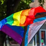 Rainbow Pride flag waving outdoors, with a transgender flag overlay: bright colors in motion near a building.