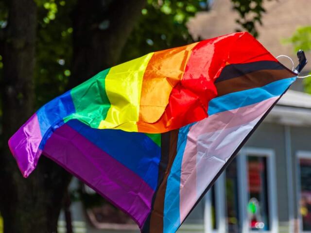 Rainbow Pride flag waving outdoors, with a transgender flag overlay: bright colors in motion near a building.