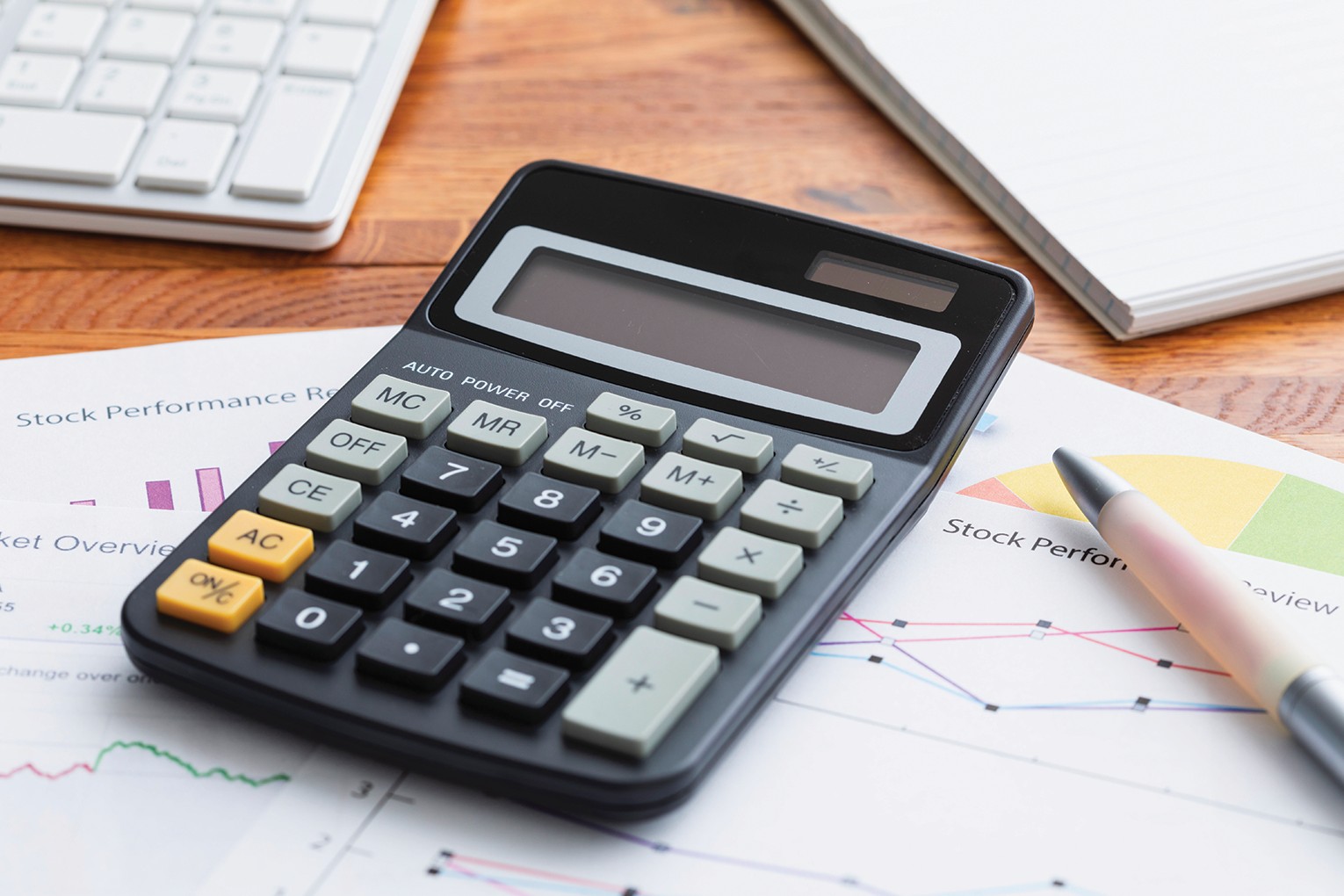 Calculator resting on stock performance reports with a pen and charts nearby on a wooden desk