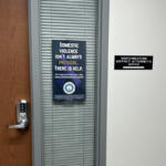 Wooden door with a silver electronic lock beside a window with blinds; poster about domestic violence on the blinds area.