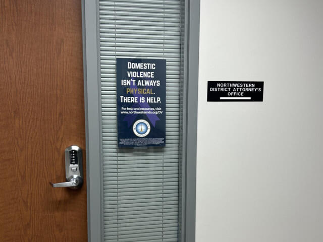 Wooden door with a silver electronic lock beside a window with blinds; poster about domestic violence on the blinds area.