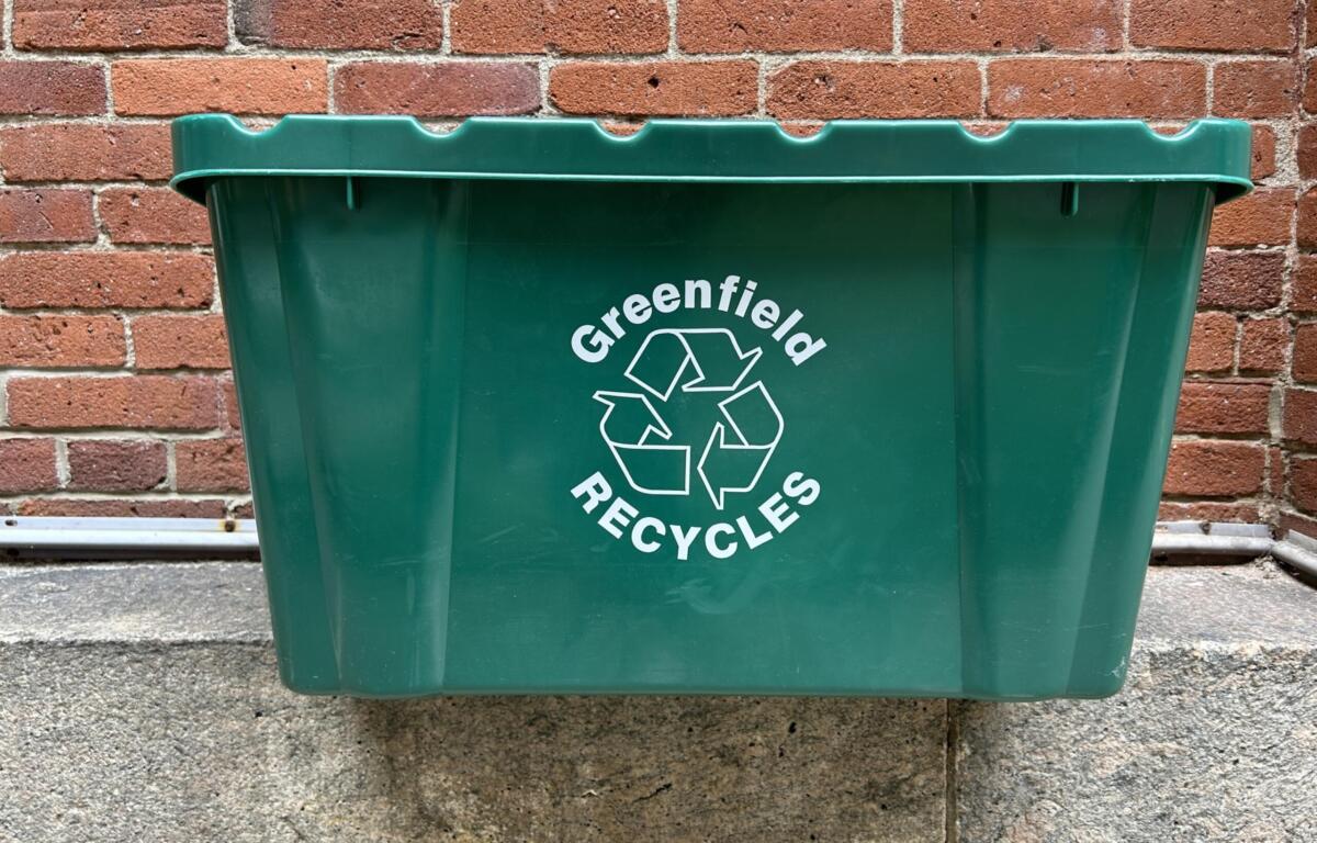 Green recycling bin in front of a red brick wall, showing a white recycling symbol and the word 'Greenfield' on the side.
