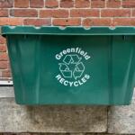 Green recycling bin in front of a red brick wall, showing a white recycling symbol and the word 'Greenfield' on the side.