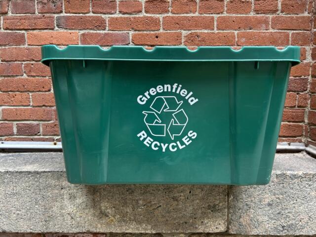 Green recycling bin in front of a red brick wall, showing a white recycling symbol and the word 'Greenfield' on the side.