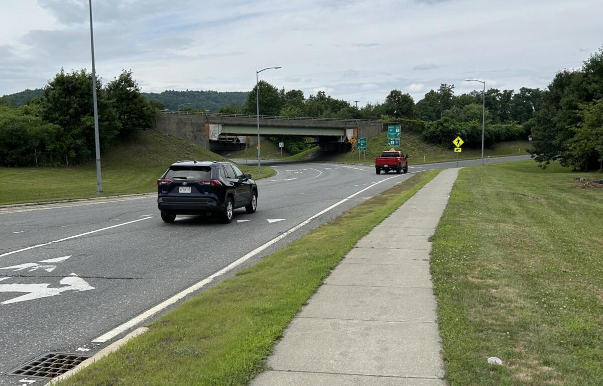 Road with a concrete underpass ahead; two cars on the street, a sidewalk and grassy verge on the right under an overcast sky.