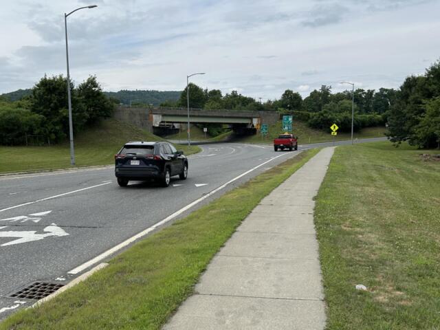 Road with a concrete underpass ahead; two cars on the street, a sidewalk and grassy verge on the right under an overcast sky.