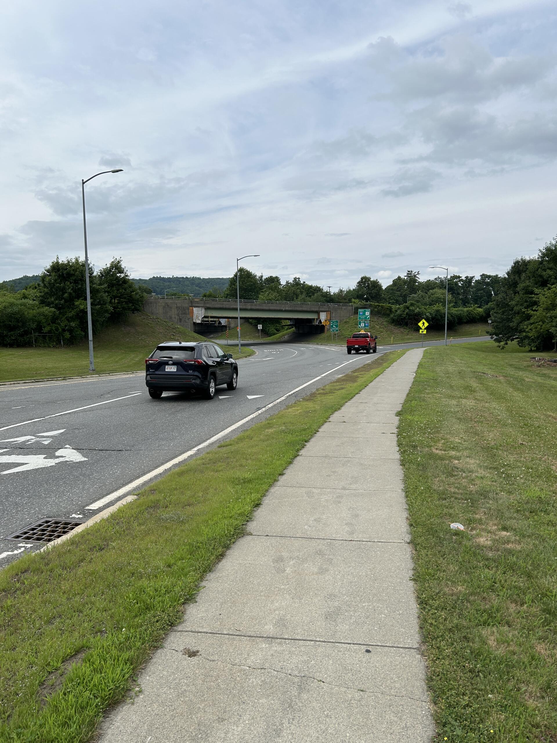 Road with a concrete underpass ahead; two cars on the street, a sidewalk and grassy verge on the right under an overcast sky.