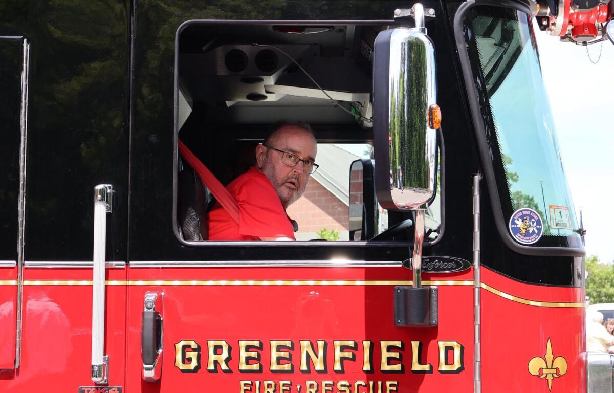 Firefighter in a red turnout jacket sits in the open cab of a black and red fire truck, looking toward the camera with glasses on.