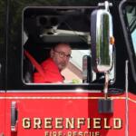 Firefighter in a red turnout jacket sits in the open cab of a black and red fire truck, looking toward the camera with glasses on.