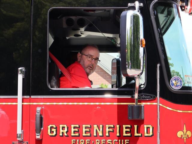 Firefighter in a red turnout jacket sits in the open cab of a black and red fire truck, looking toward the camera with glasses on.