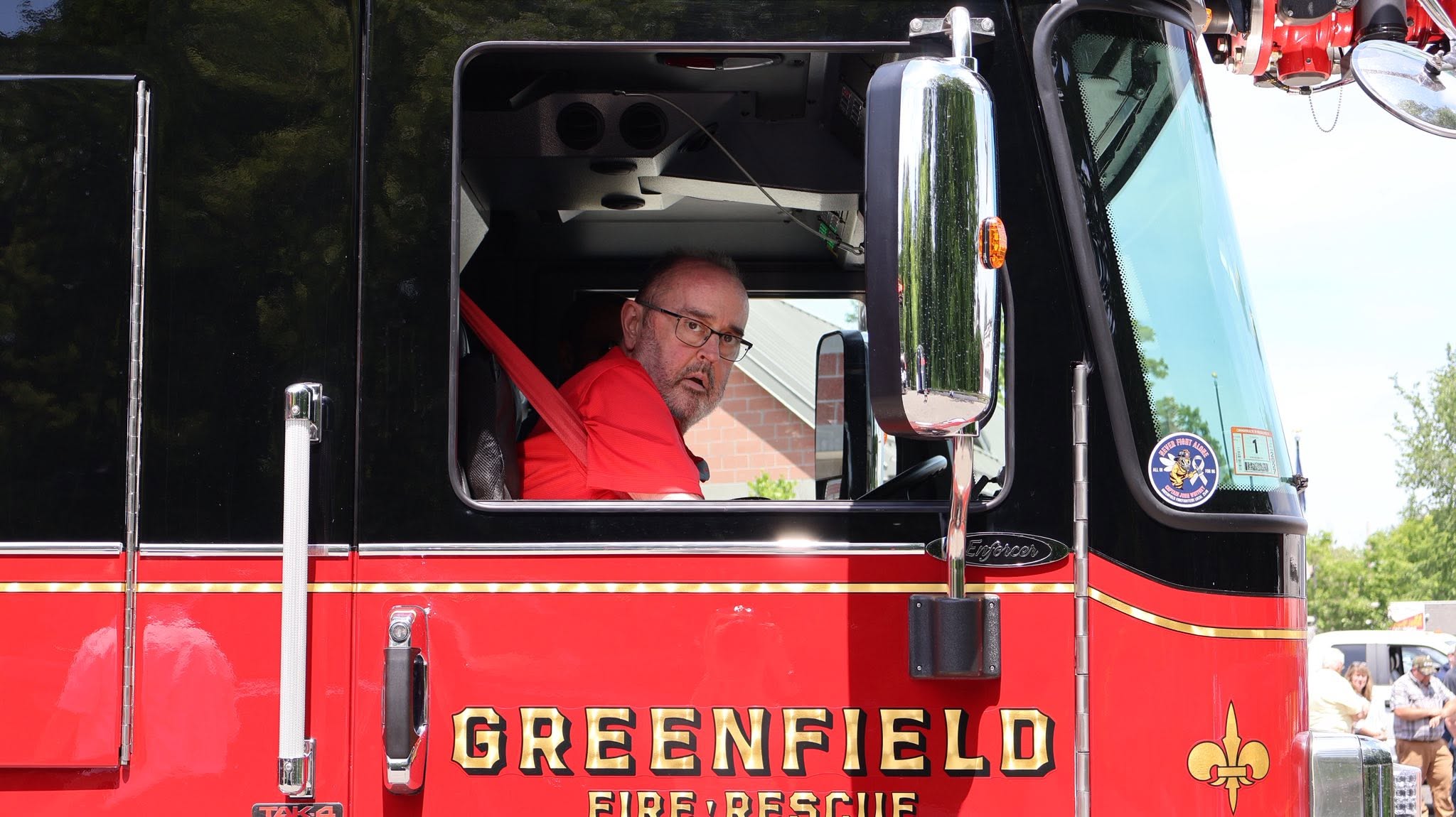 Firefighter in a red turnout jacket sits in the open cab of a black and red fire truck, looking toward the camera with glasses on.