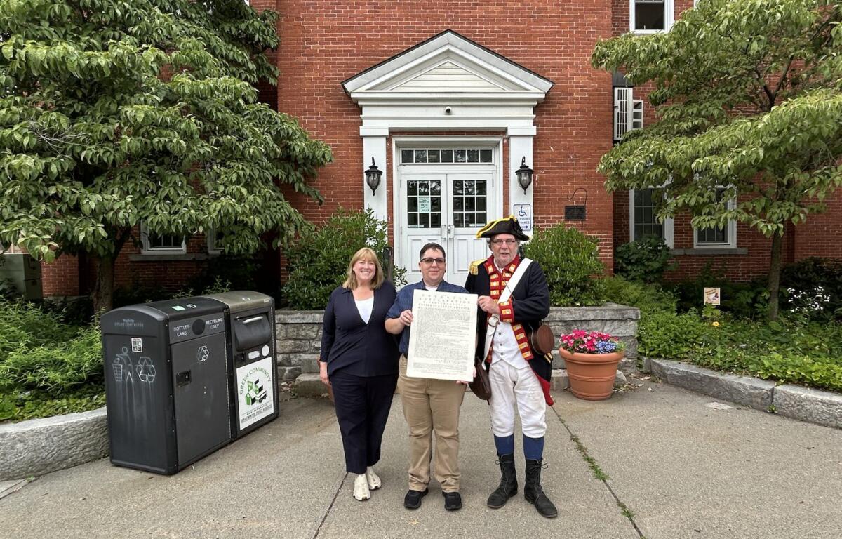 Three people stand outside a brick building holding a large document, with a costumed colonial-era figure on the right.