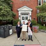 Three people stand outside a brick building holding a large document, with a costumed colonial-era figure on the right.